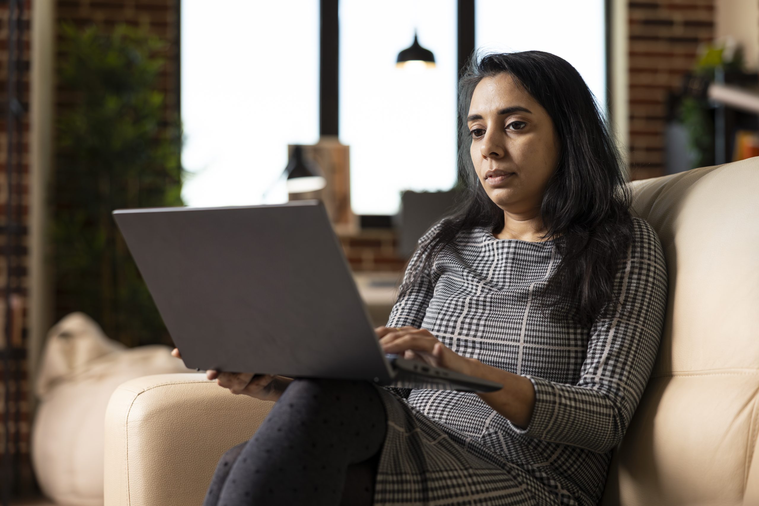 Modern Indian woman seated on couch, focused on editing blog post and researching business trends. Female entrepreneur working remotely with laptop, reviews marketing plans in cozy home environment.